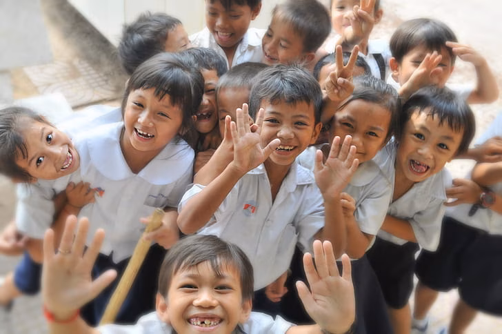 A group of school children wearing white uniforms gather around the camera smiling, giving peace signs and holding their hands in the air