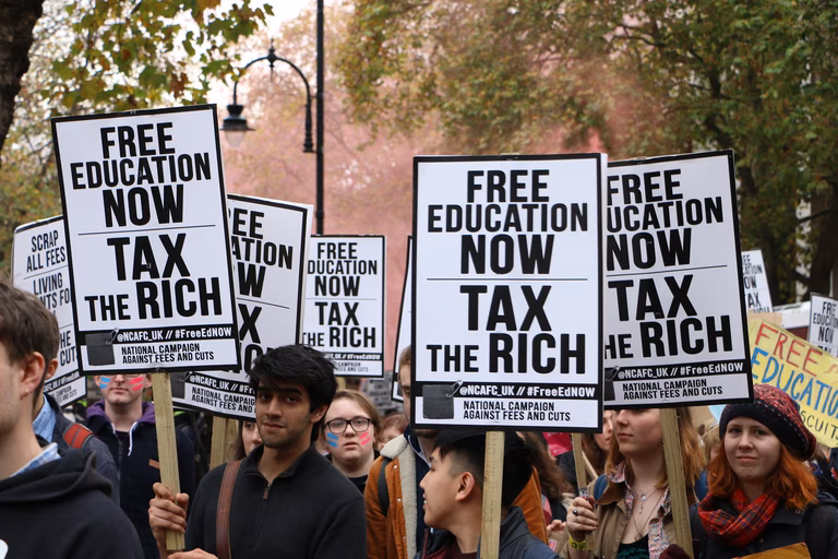 Protesters stand in a green park holding signs calling for free education and a tax on the rich
