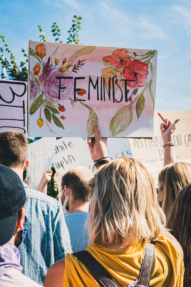 View from the back of a women's rights protest, there is a large white poster that says 'Feminist' with beautiful flowers painted in the background