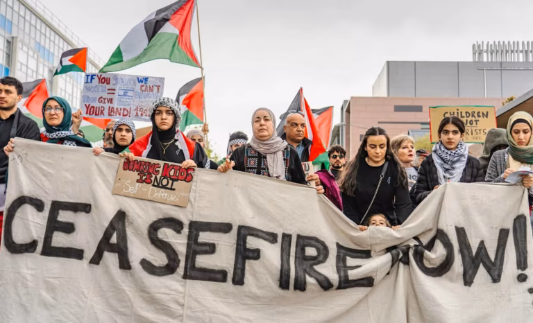 Protesters stand in the street calling for an end to the fighting in Gaza. They hold a large white banner which reads 'Ceasefire Now'