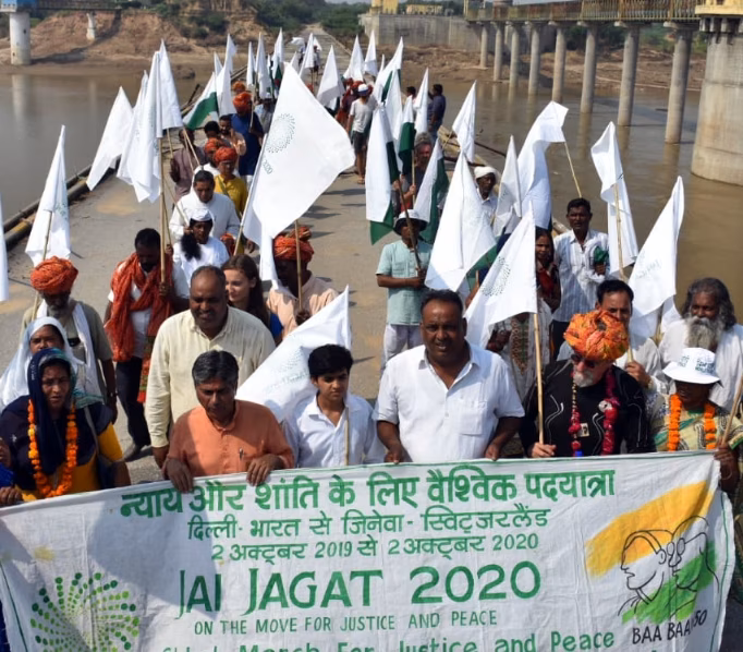 Jai Jagat 2020 crossing the Chambel river, Madhya Pradesh, India. Many people walk across a bridge holding white flags and a large banner with the organisations name on.