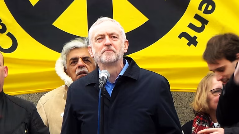 Jeremy Corbyn speaking at the #StopTrident rally at Trafalgar Square in February 2016. He stands infront of a large yellow CND poster.
