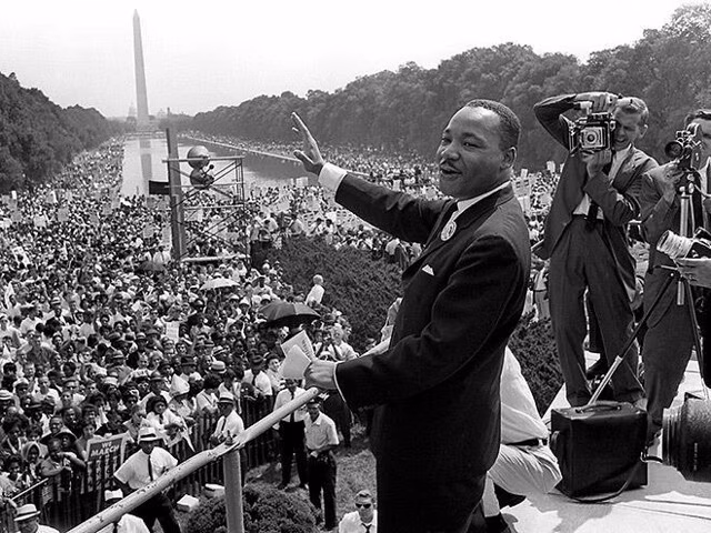 Black and white image of Martin Luher King Jr standing above a huge crowd delivering a speech.