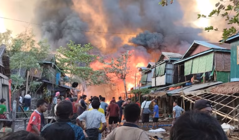 MAF bombing Kyauktaw Township in Myanmar as part of their civil war. A large explosion and cloud of smoke fill the sky at the end of a street of wodden houses. Many onlookers standby. 