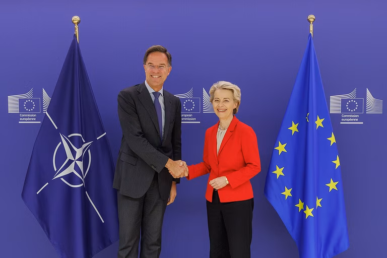 NATO Secretary General Mark Rutte with President of the European Commission Ursula von der Leyen in Brussels, 29 October 2024. They stand shaking hands looking into the camera infont of the NATO and EU flags.