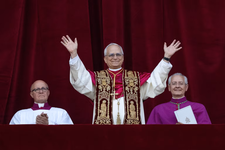 Pope Leo XIV addresses an audience with open hands, he is wearing white and purple robes and is smiling warmly.