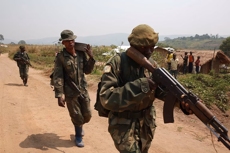 Three soldiers walk through a village in the DRC carrying weapons. Villagers stand closeby in the background. 