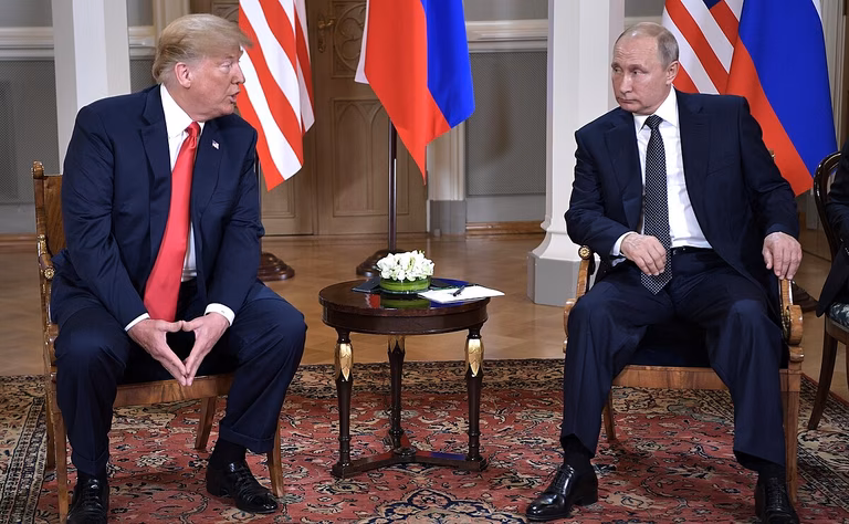 Vladimir Putin & Donald Trump at a meeting in Helsinki. They sit next to a small table with their respective flags in the background.