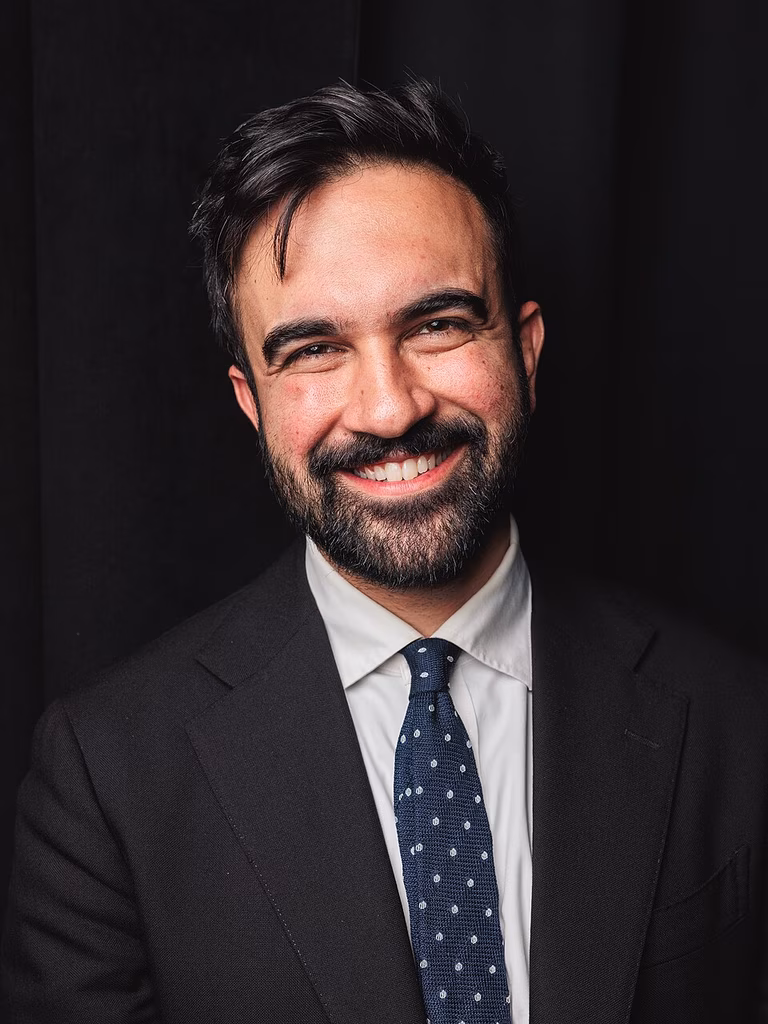 Zohran Mamdani wears a black suit and white shirt and smiles warmly at the camera infront of a black background.